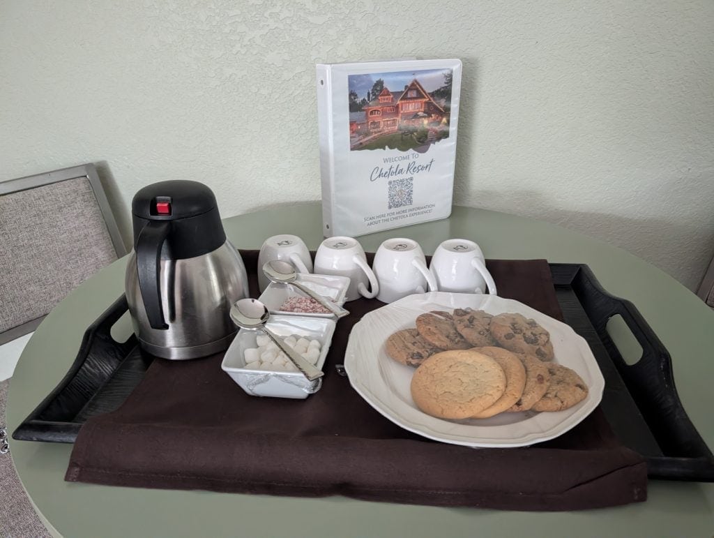 A welcoming tray in a room at the Chetola Resort in Blowing Rock, NC, featuring a plate of assorted cookies, a thermos of hot cocoa, white mugs, and small dishes with marshmallows and peppermint pieces, accompanied by an informational pamphlet about the resort.