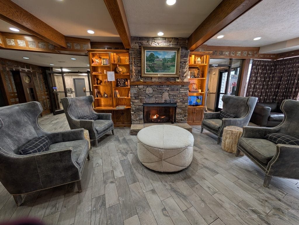 A cozy lobby at Chetola Lodge in Blowing Rock, featuring a stone fireplace with a framed painting above it, surrounded by plush armchairs, a circular ottoman, and wooden bookshelves displaying books and decor.