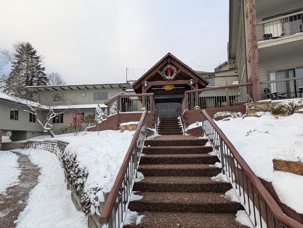 Snow-covered entrance to the Chetola Resort in Blowing Rock, NC, featuring a central staircase with wooden railings leading to a rustic lodge-style building adorned with a holiday wreath and festive decorations.