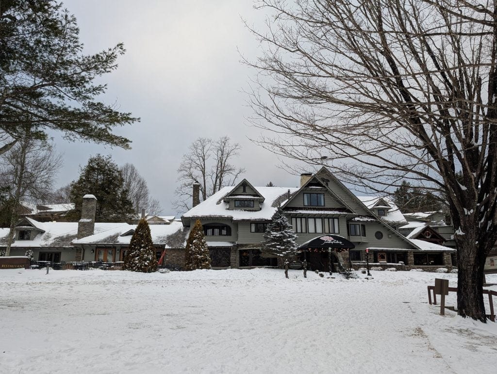 A snowy exterior view of Timberlake's Restaurant at the Chetola Resort, featuring a charming lodge-style building with a steep roof, stone chimneys, and festive decorations, surrounded by bare winter trees.