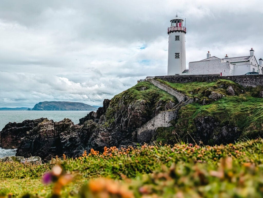 Fanad Head Lighthouse perched atop rugged cliffs in Donegal, Ireland, with stone steps leading up through green hills and the Atlantic Ocean stretching out under dramatic skies in the background.