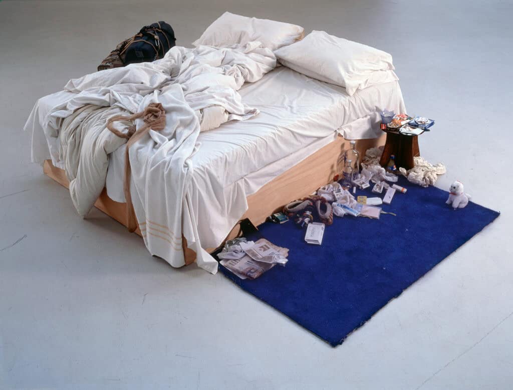 A wide shot of Tracey Emin's iconic contemporary art installation My Bed, featuring an unmade bed surrounded by personal detritus on a blue rug.