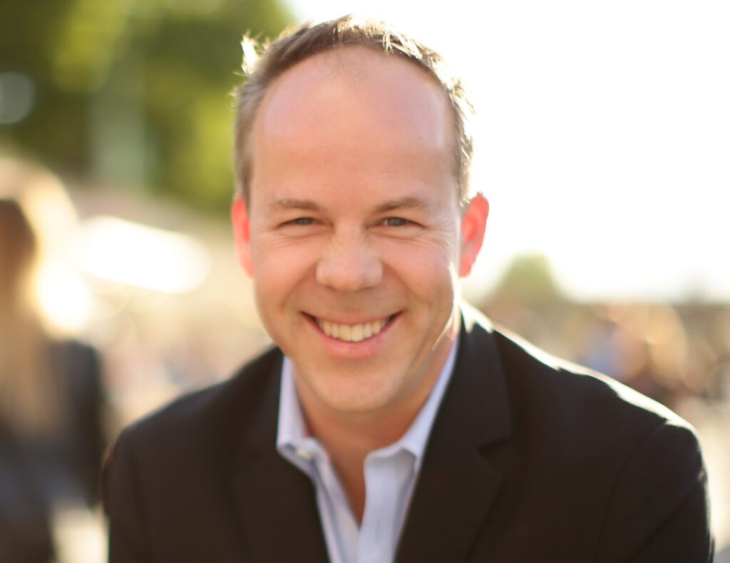 Derek Fredrickson, founder and CEO of The COO Solution is sitting outside on a sunny day wearing a suit while smiling and looking at the camera