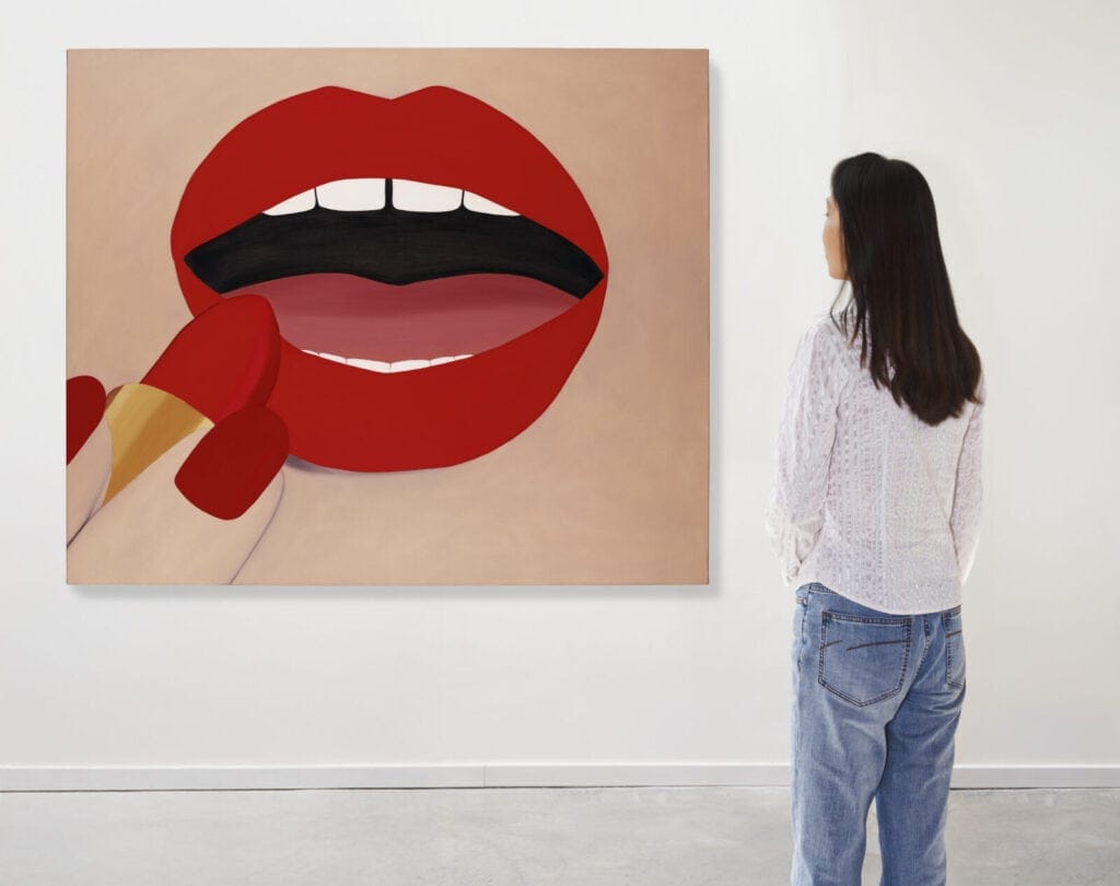 A woman in casual clothes stands in a gallery, looking at a Lize Bertelli's painting showing a red lips being colored with red lipstick on a beige background.