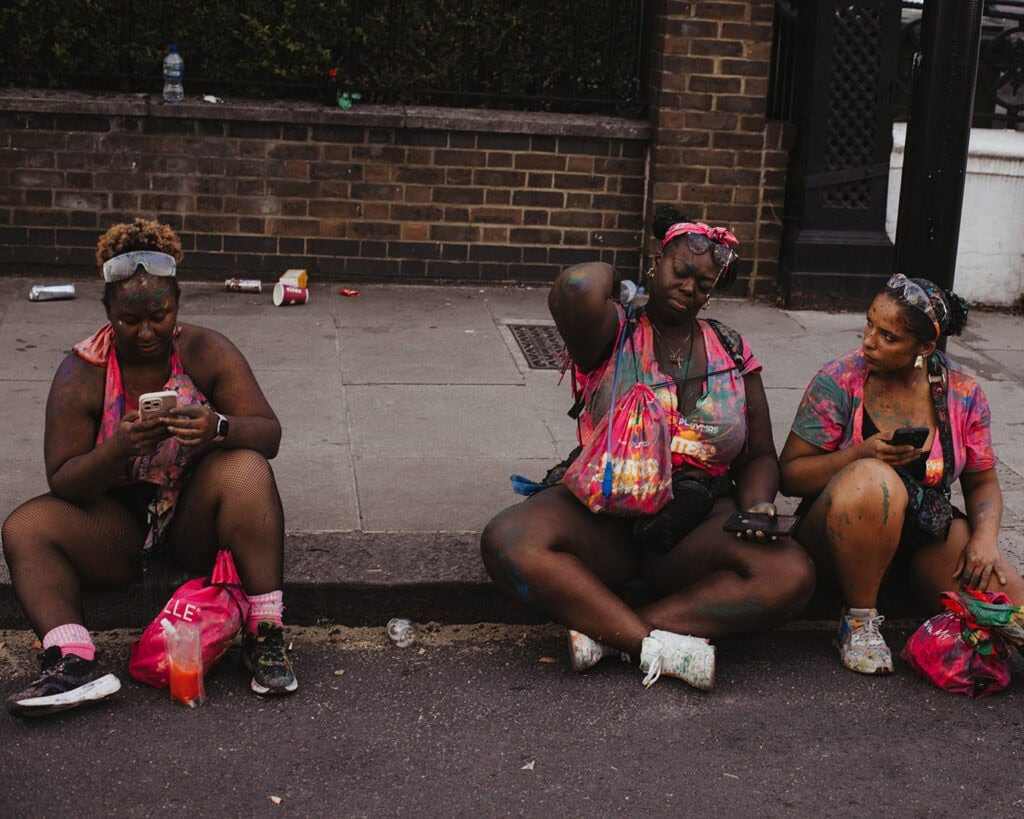 Carnival-goers resting on the curb covered in paint after J’Ouvert at Notting Hill Carnival.