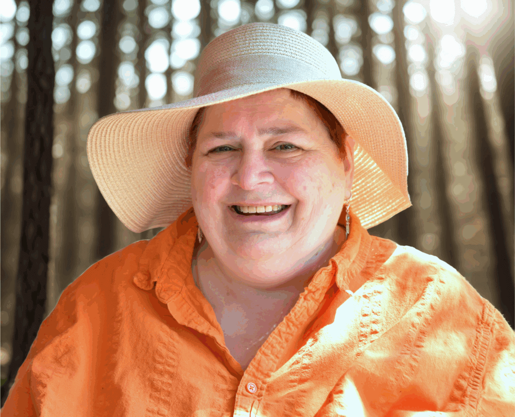 Bild von mir mit orangener Bluse und großem Sonnenhut, wie immer grinsend