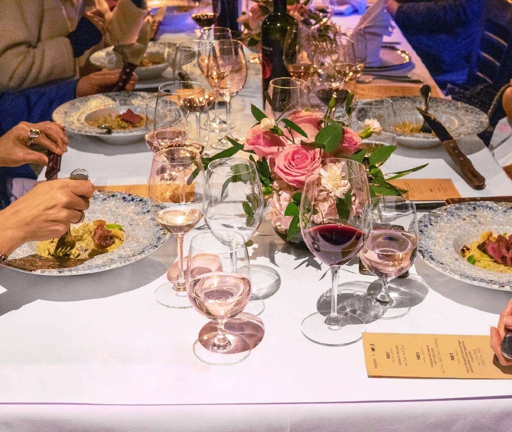 Mesa servida con copas de vino, flores y plato de carne con risotto durante una cena en La Vaca Loca