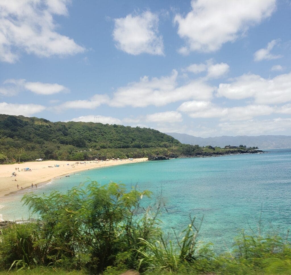 A beautiful beach in Oahu with golden sand, clear turquoise water, and lush green hills in the background. The beach is dotted with visitors enjoying the sunny day under a partly cloudy sky.