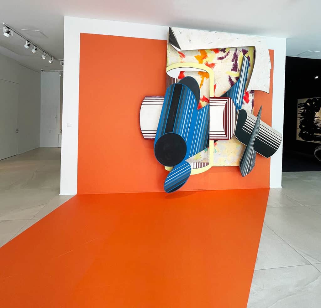A bold abstract wall sculpture featuring colorful, striped, and geometric shapes extends from an orange wall—an eye-catching piece reminiscent of Miami Art Week 2025, with the vibrant orange color flowing seamlessly across the floor.