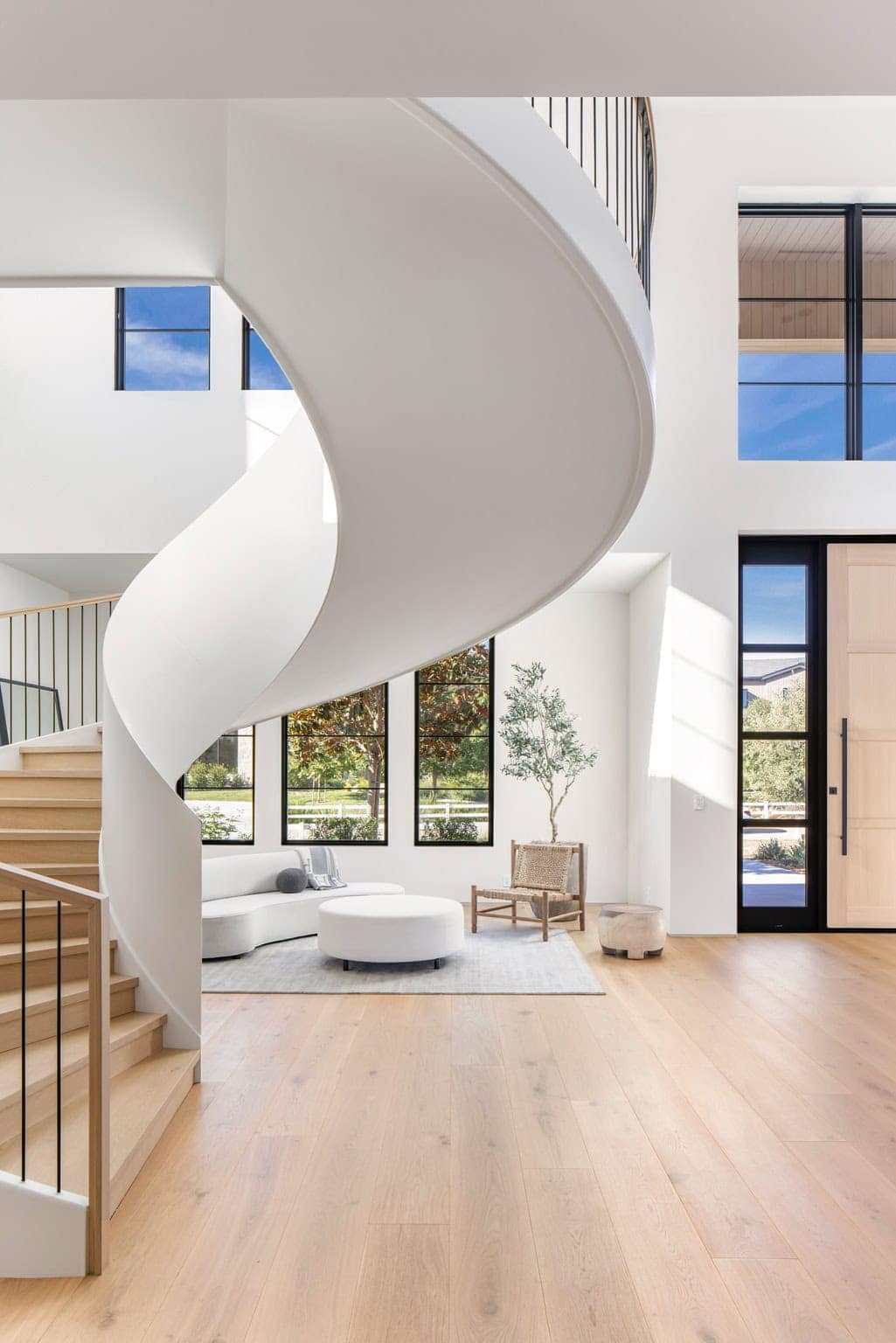 Contemporary minimalist interior featuring a curved white staircase, large black-framed windows, natural wood flooring, and soft neutral furnishings.