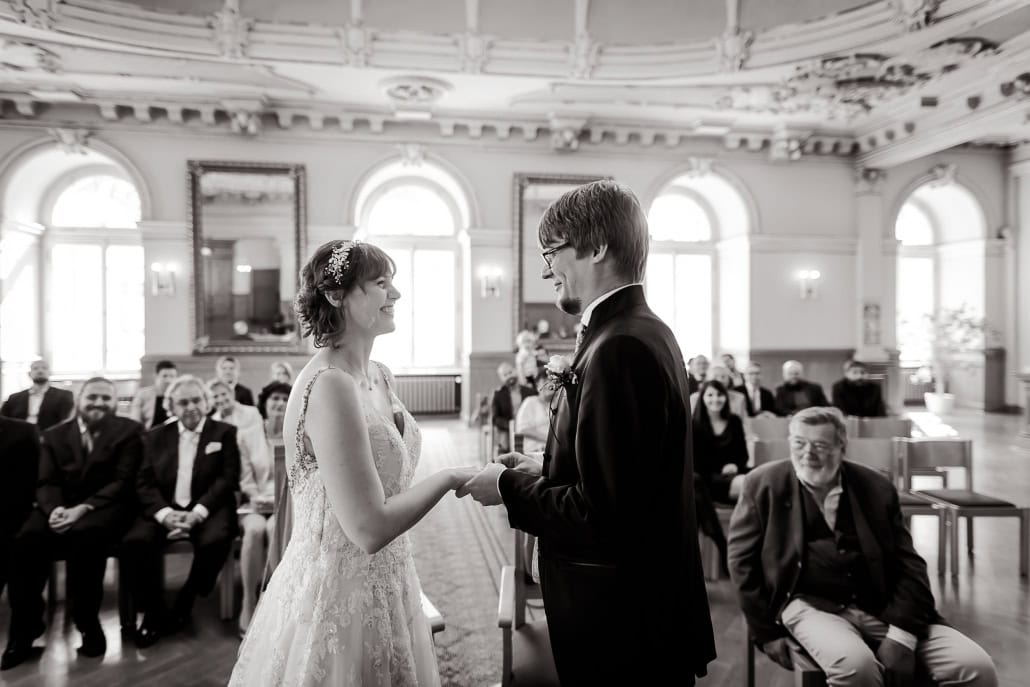 Ein Augenblick inniger Verbundenheit: das Hochzeitspaar hält sich liebevoll die Hände und tauscht verliebte Blicke und die Ringe in einem feierlichen Moment. Umgeben von den Zeugen ihrer Liebe, versprechen sie sich eine gemeinsame Zukunft voller Romantik und Glück