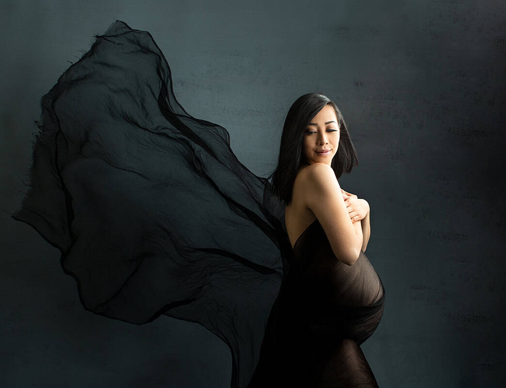 Elegant gravidfotografering av en gravid kvinne i svart bandagenskjørt med flytende stoff rundt skuldrene, som fremhever morsliv og skjønnhet, mot en enkel mørk bakgrunn.