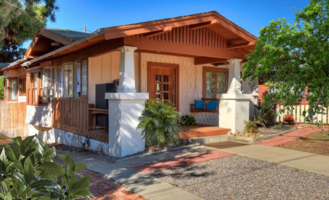 Exterior side view of the historic Craftsman bungalow at 1930 Granada Avenue in South Park with a covered front porch, white pedestal columns, and a wooden porch swing under a clear blue sky.