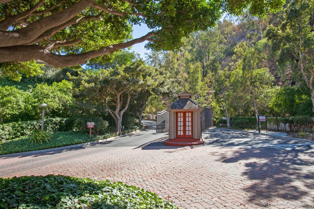 Gated community entrance at 5474 Caminito Borde featuring a brick-paved driveway, a security kiosk with red doors, and lush green hillside landscaping.