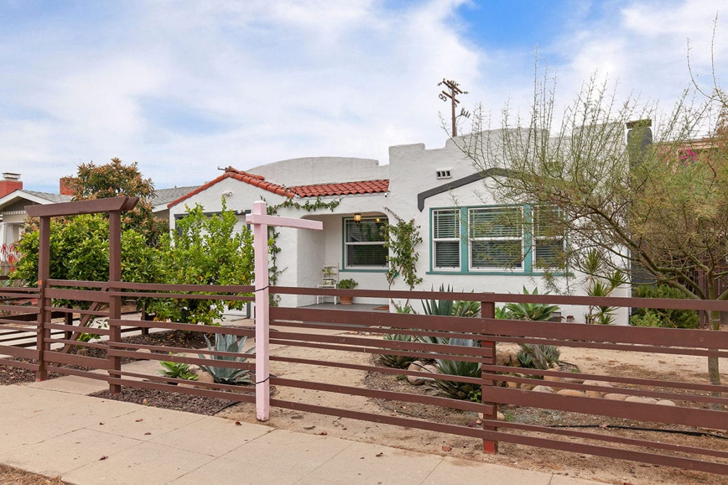 Spanish-style home at 4243 46th Street in City Heights featuring a white stucco exterior, red clay roof tiles, and a horizontal wood fence with a pink sign post.