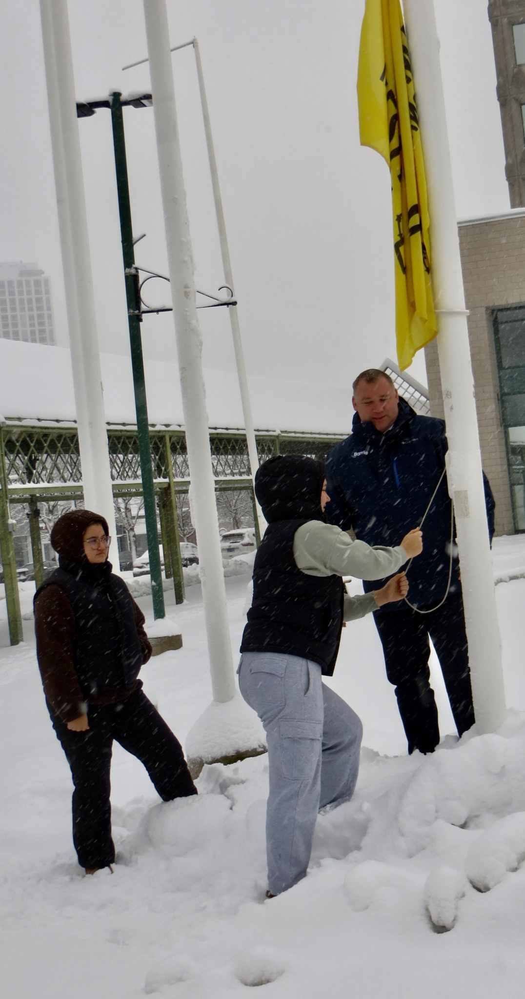“Three people standing in deep snow, raising a yellow Amnesty International flag on a flagpole during a winter Write for Rights community action.”