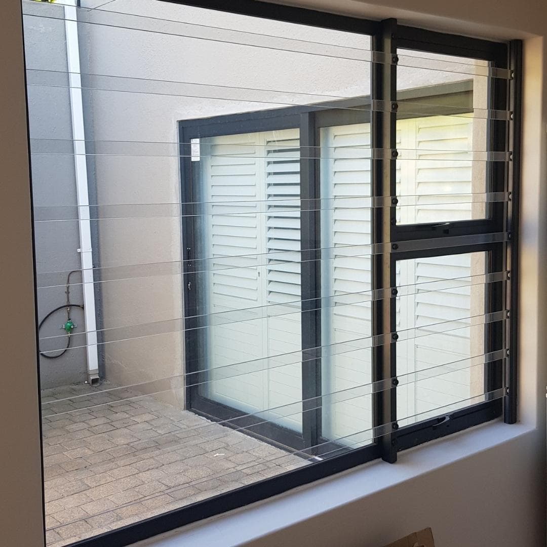 Modern black-framed window fitted with clear bars burglar bars, its see-through security strips spanning a paved courtyard outlook.