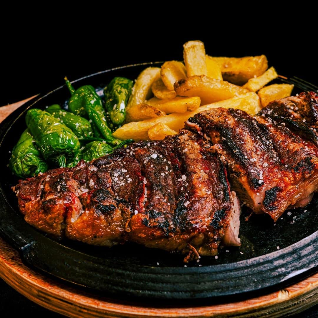 A plate of medium rare, charred flat iron steak with a side of hand cut fries and roasted peppers.