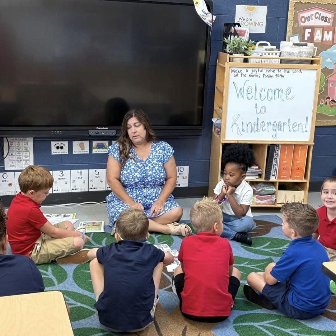Kindergarten teacher Mrs. Trish Hull sits on the classroom rug with her kindergartners gathered in a circle at Martin Luther School, Joplin MO, beside a Welcome to Kindergarten sign