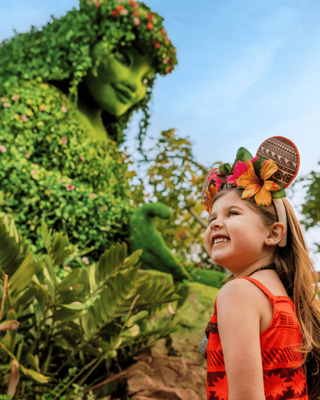 Young girl smiling at a green Disney character in a garden setting.