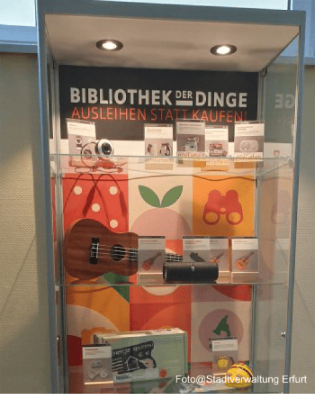 Eine Galsvitrine der Bibliothek der Dinge in Erfurt, die auf unterschiedlichen Ebenen mit ausleihbaren Sachen bestückt sind. Darunter unter anderem eine Ukulele, eine Webcam, ein Spielzeug.