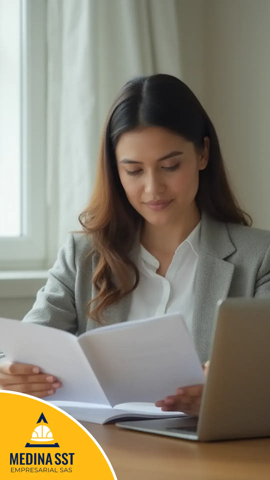 Profesional sentada a un escritorio lee un manual impreso abierto entre las manos, junto a un computador portátil