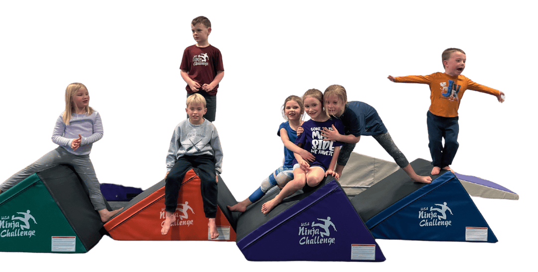 Seven happy children sitting and standing on colorful triangular wedge mats branded with the USA Ninja Challenge logo.