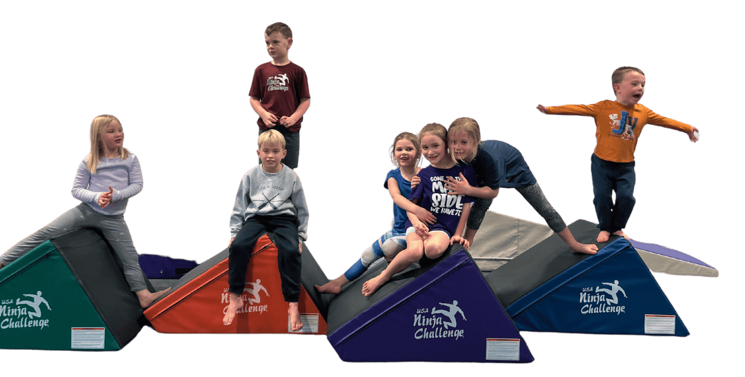 Seven happy children sitting and standing on colorful triangular wedge mats branded with the USA Ninja Challenge logo.