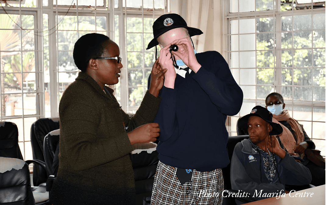 A girl with albinism holding a monocular close to her eye while a lady is standing next to her.