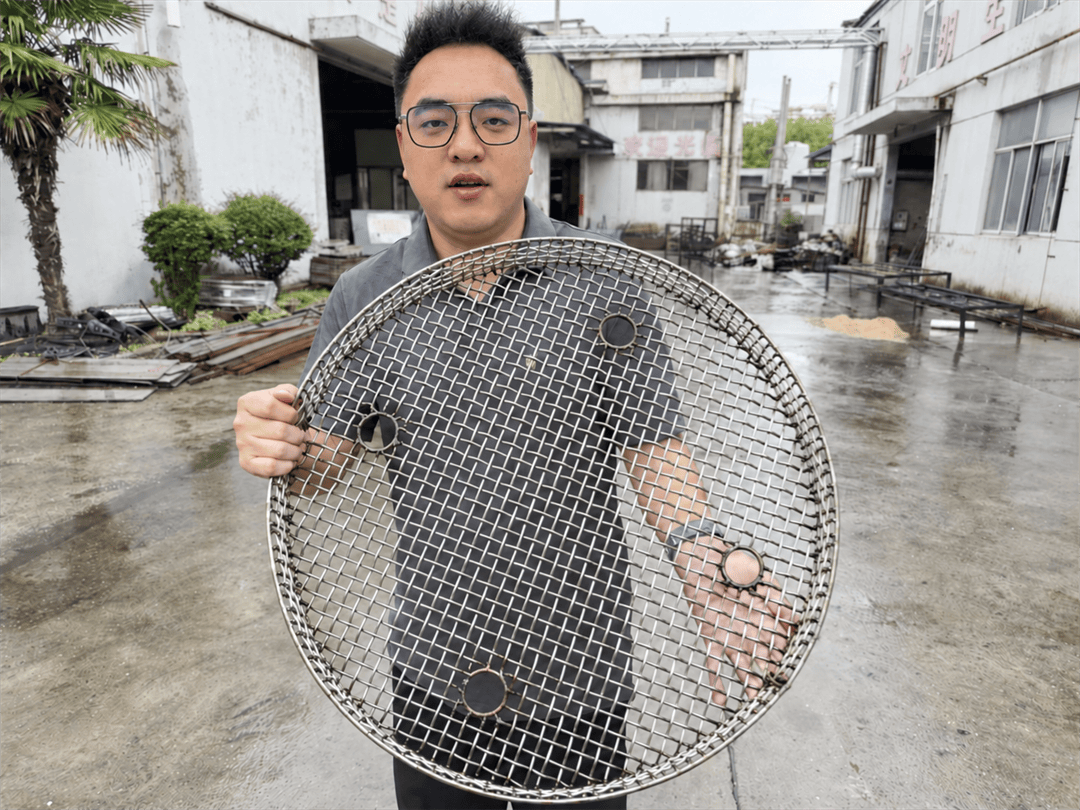 Jonny holding a round wire-mesh furnace charging basket in the ECOOSUN factory courtyard