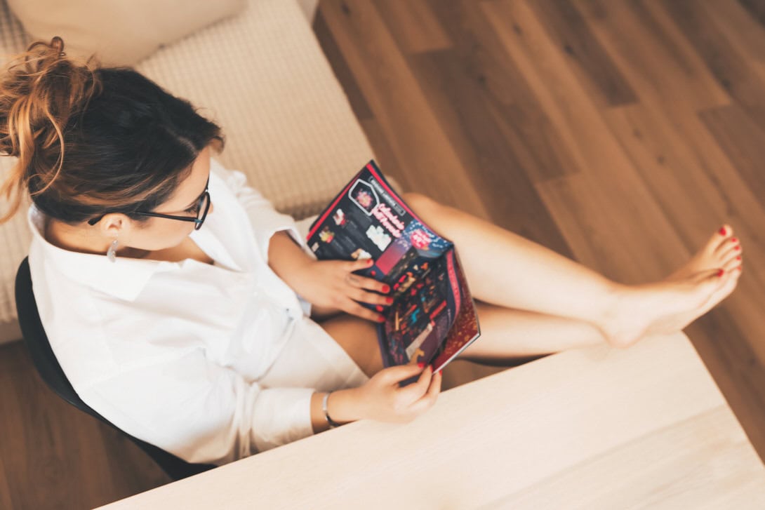 Donna che legge una rivista mentre è seduta su un divano, indossando una camicia bianca e con le gambe incrociate. L'ambiente è luminoso e accogliente, con pavimento in legno.