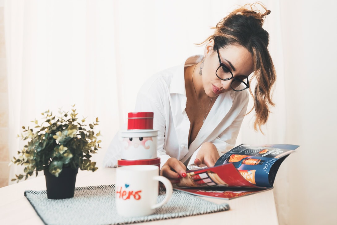 Donna con occhiali che legge una rivista su un tavolo, con una tazza di caffè e decorazioni natalizie in primo piano.