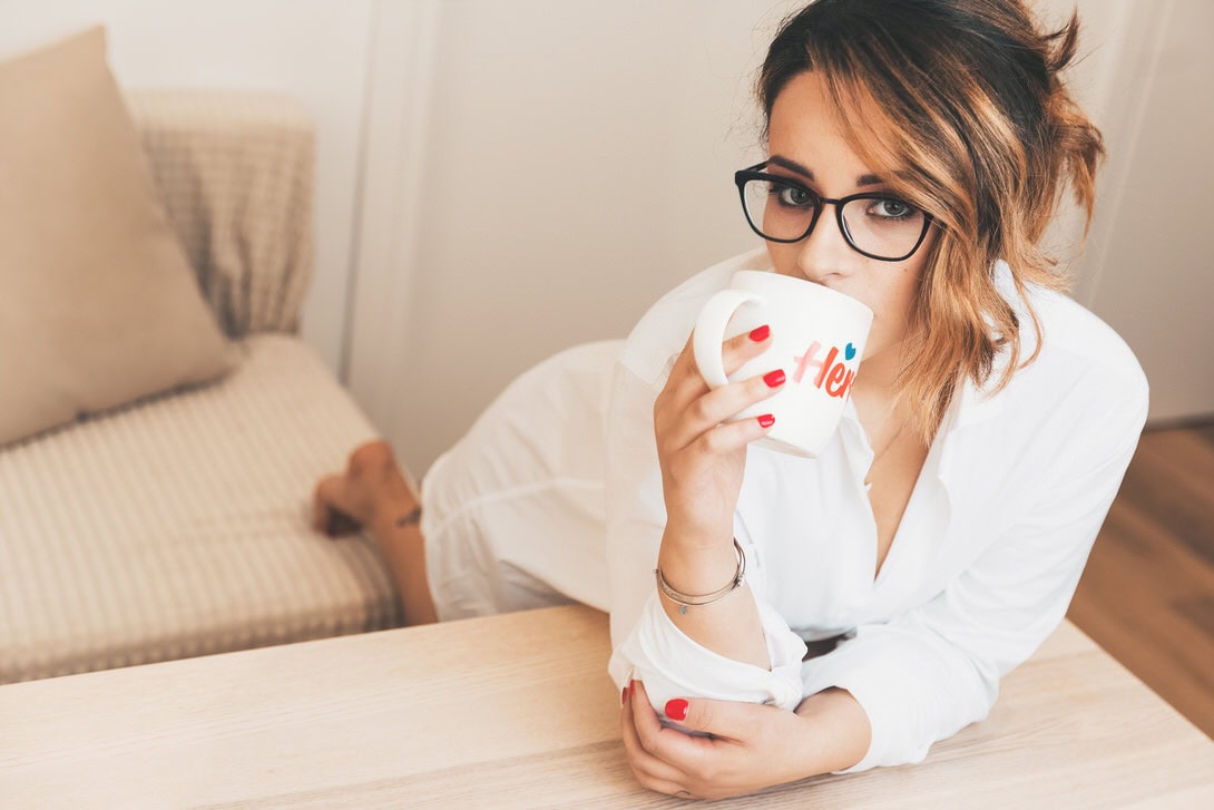 Donna in camicia bianca che sorseggia una tazza di caffè mentre si trova sul divano, con occhiali e un'espressione rilassata.