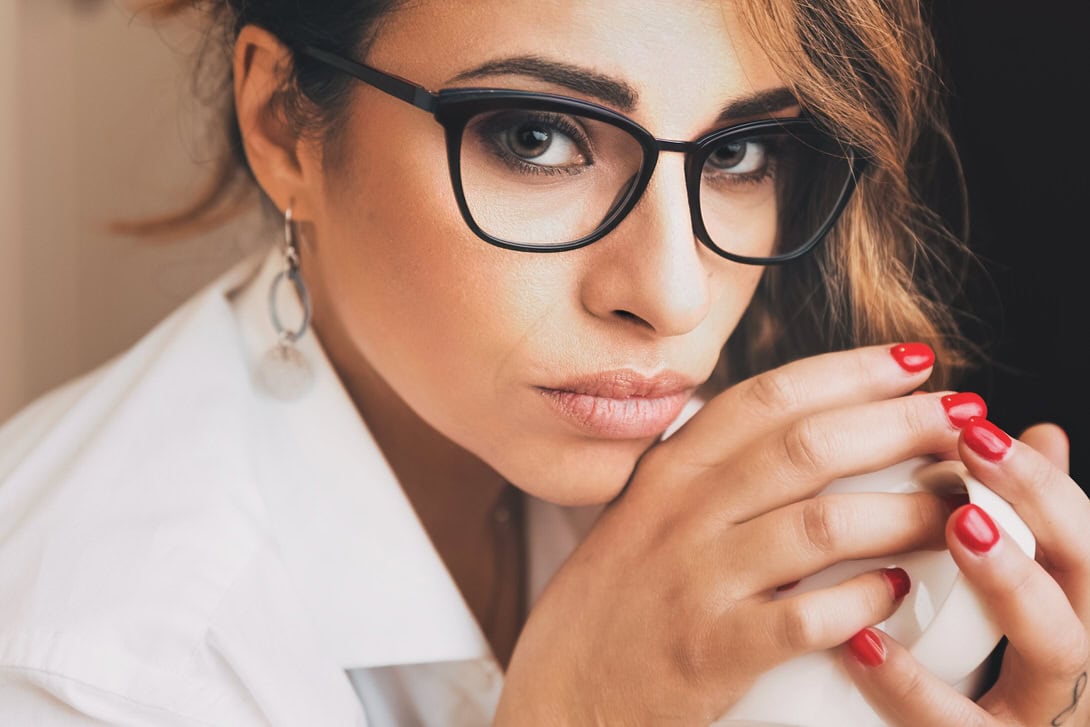 Donna con occhiali neri che tiene una tazza, con uno sguardo intenso e un'espressione pensierosa, indossando una camicia bianca.