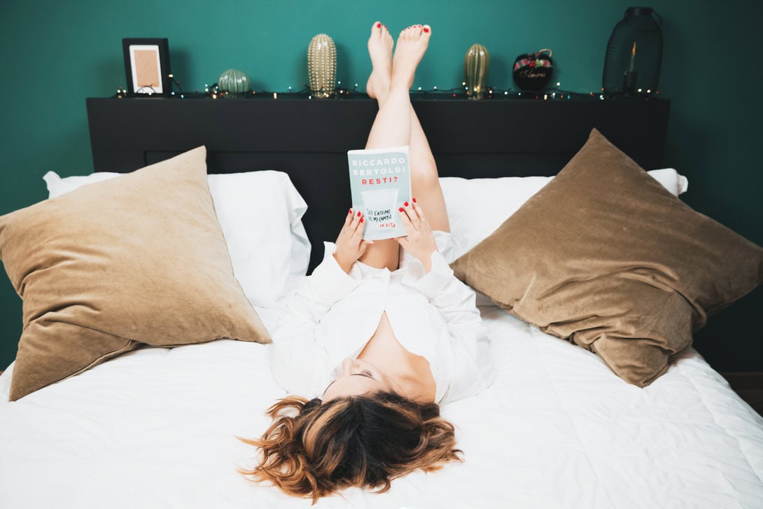 Donna sdraiata sul letto, con le gambe sollevate e un libro tra le mani, in un ambiente accogliente e rilassato. Decorazioni moderne sullo sfondo e cuscini eleganti.