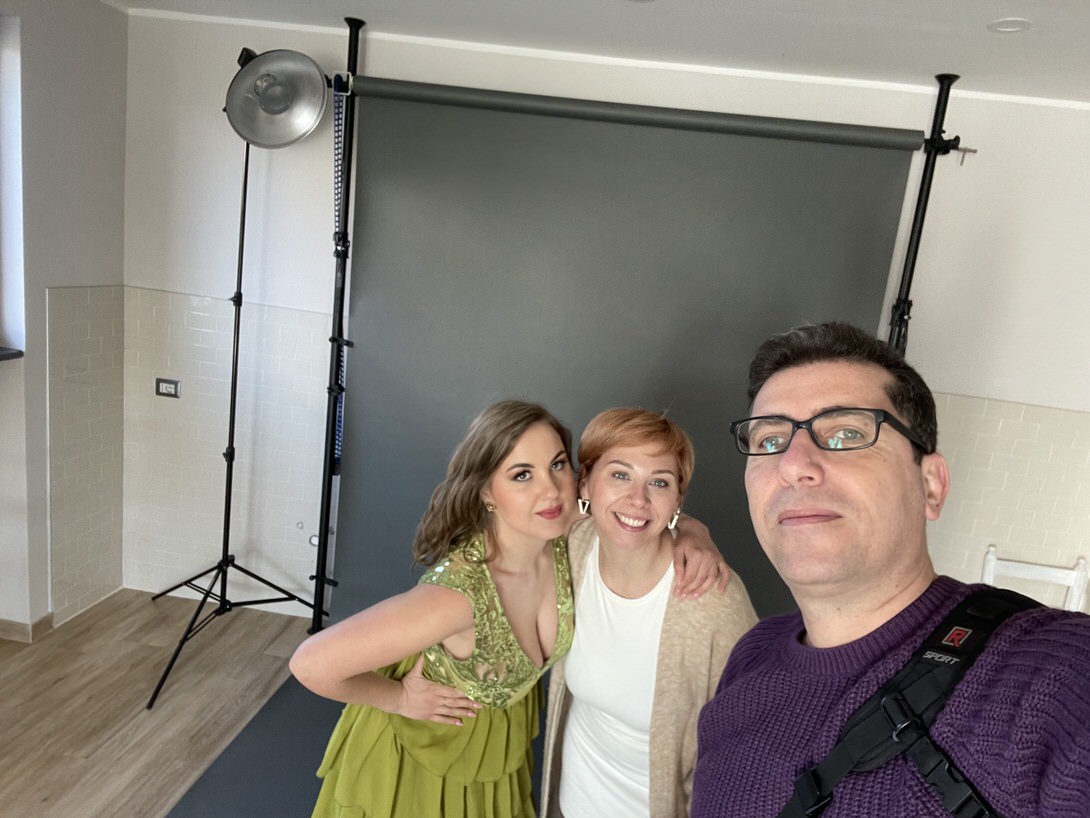 Tre persone sorridenti in uno studio fotografico, con uno sfondo grigio e attrezzatura da studio visibile. Due donne indossano abiti eleganti, mentre un uomo scatta un selfie con loro. L'ambiente è luminoso e moderno.