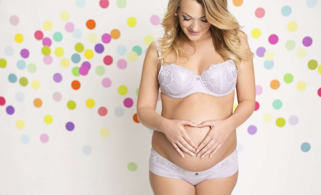 Kvinne i vakker graviditetspose som smiler og holder hendene på magen, omgitt av fargerike dots i bakgrunnen, symboliserer graviditet og mammatid life.