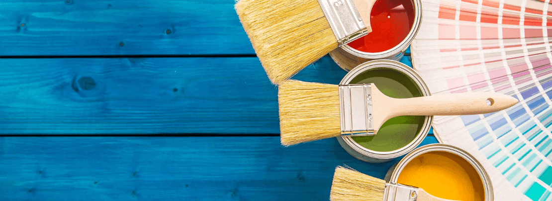 Pinsel mit roter, grüner und gelber Farbe auf blauer Holzoberfläche, daneben ein Fächer mit Farbmusterkarten für kreative Farb Ideen in der Wohnung.