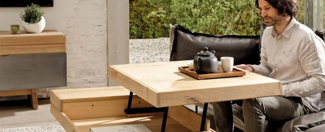 Mann sitzt auf einem Sofa vor einem höhenverstellbaren Couchtisch aus Holz mit Teeservice, der zusätzlichen Stauraum bietet. Symbolbild für Platzsparende Möbel