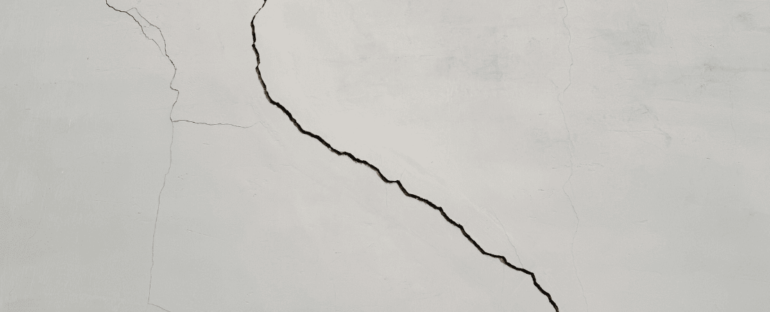 Großer Riss in einer weißen Betonwand. Die unregelmäßige Struktur des Risses zieht sich diagonal über die Oberfläche und symbolisiert Bauschäden, Materialermüdung oder strukturelle Probleme.