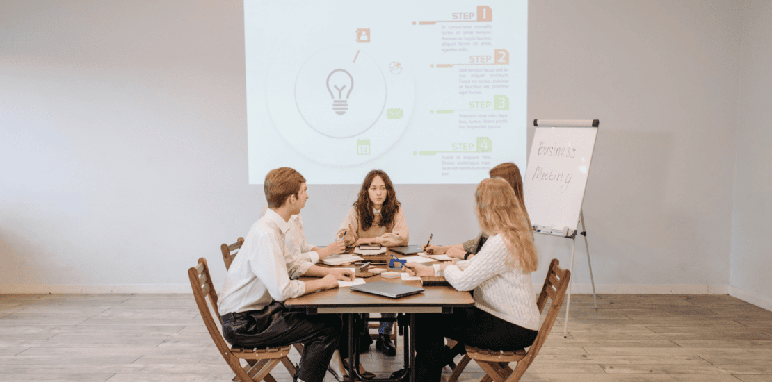 Marketing colleagues sitting in front of a desk together with a projector behind them projecting marketing steps for sales about gender equality in their business