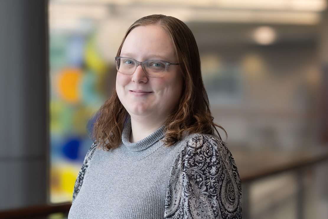 Woman with blue eyes and glasses and shoulder-length light brown hair. Wearing a gray sweater with patterned sleeves. Blurred colorful background.