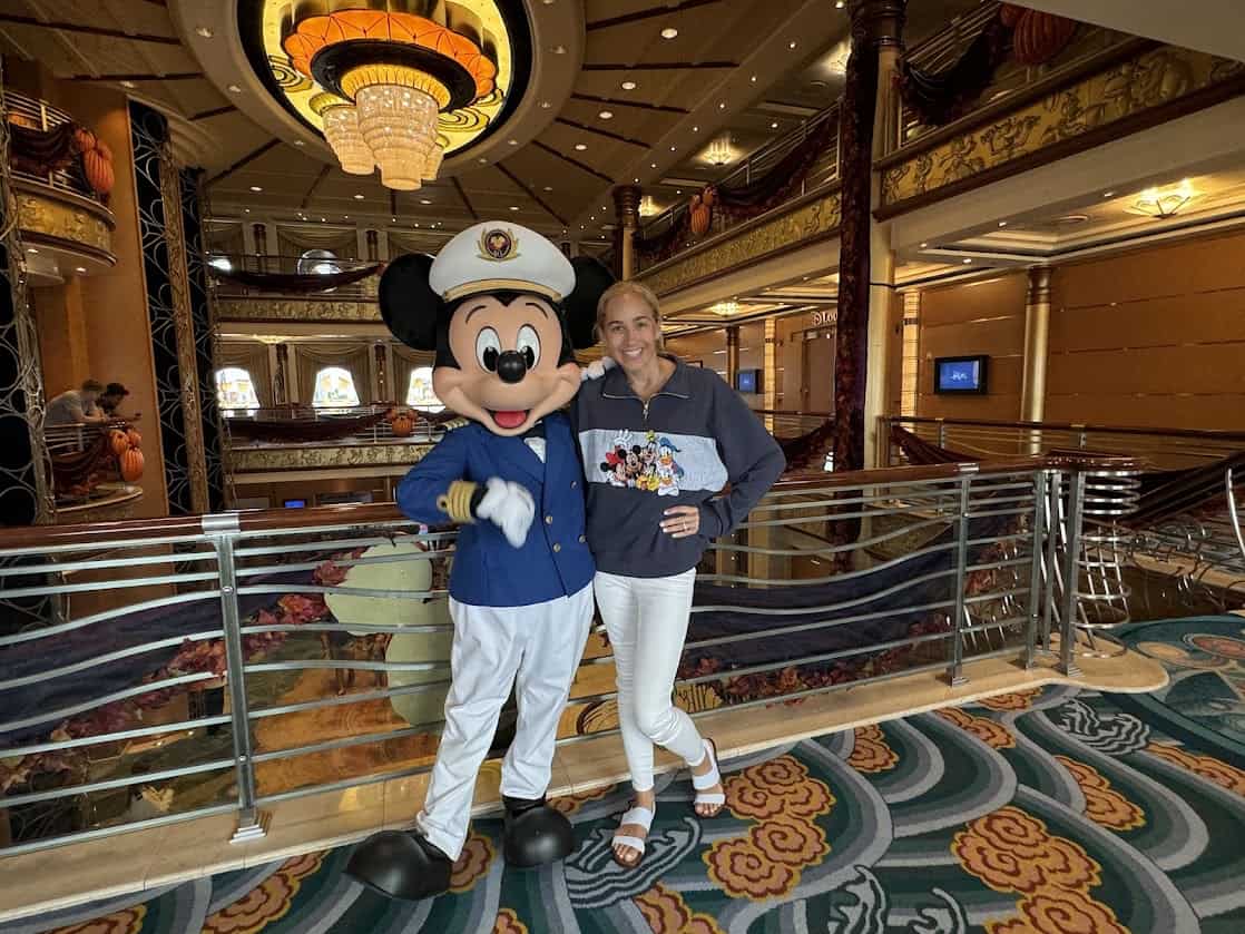Mickey Mouse dressed as a captain and a woman smiling inside a luxurious Disney cruise ship lobby.
