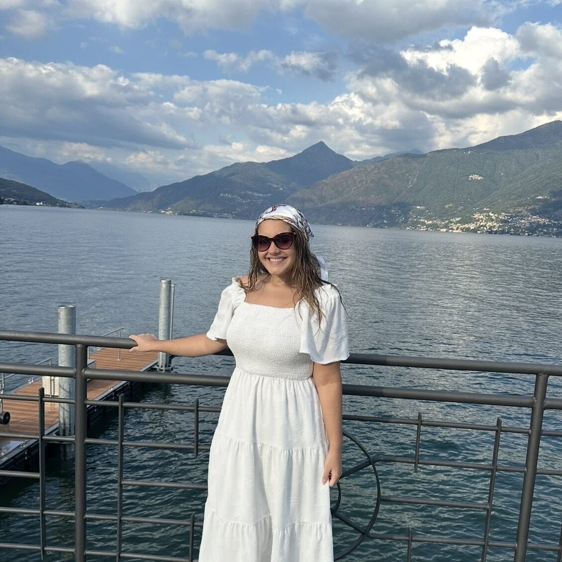 Woman in white dress enjoying lake view with mountains and sky in background.