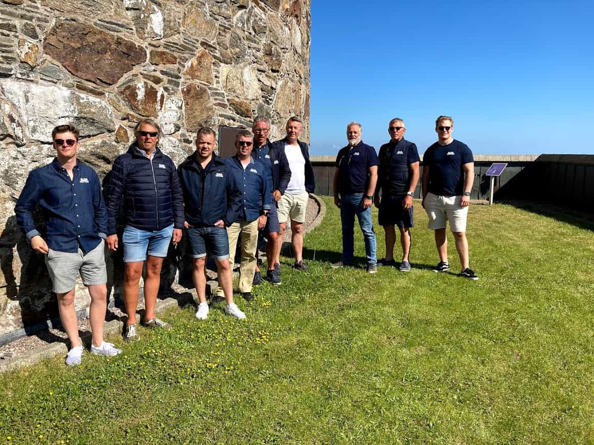 Gruppbild med åtta personer utomhus framför ett gammalt stenvägg, klädda i marinblåa arbets- eller företagskläder, på en solig dag vid vatten eller kust.