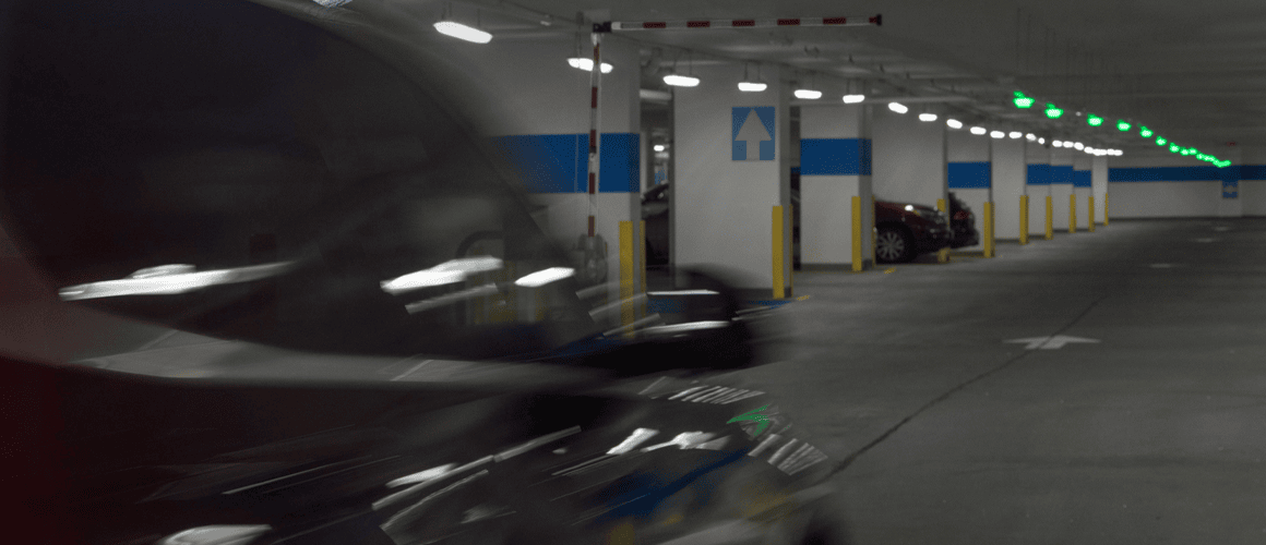 Vehicle passing under a gate into a well-lit parking garage with vacant spots and green overhead M5 smart sensors.