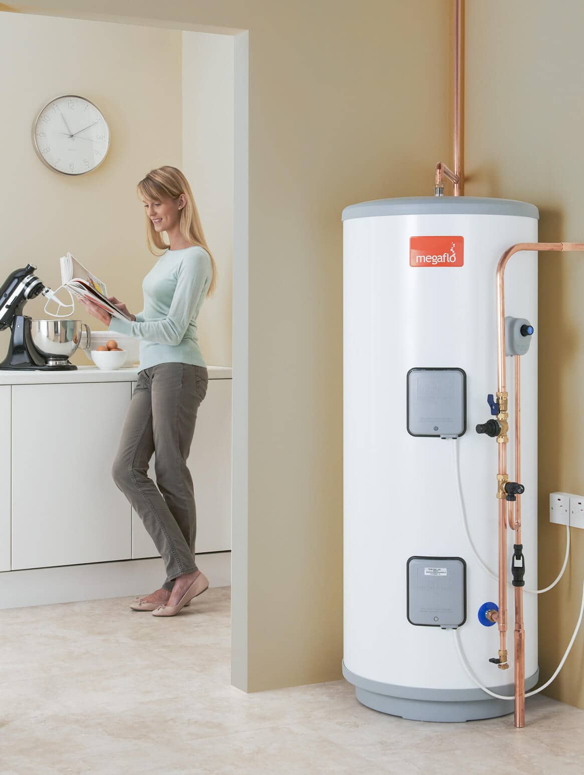 Megaflo unvented hot water cylinder in home setting. Lady smiling to left reading a cook book.
