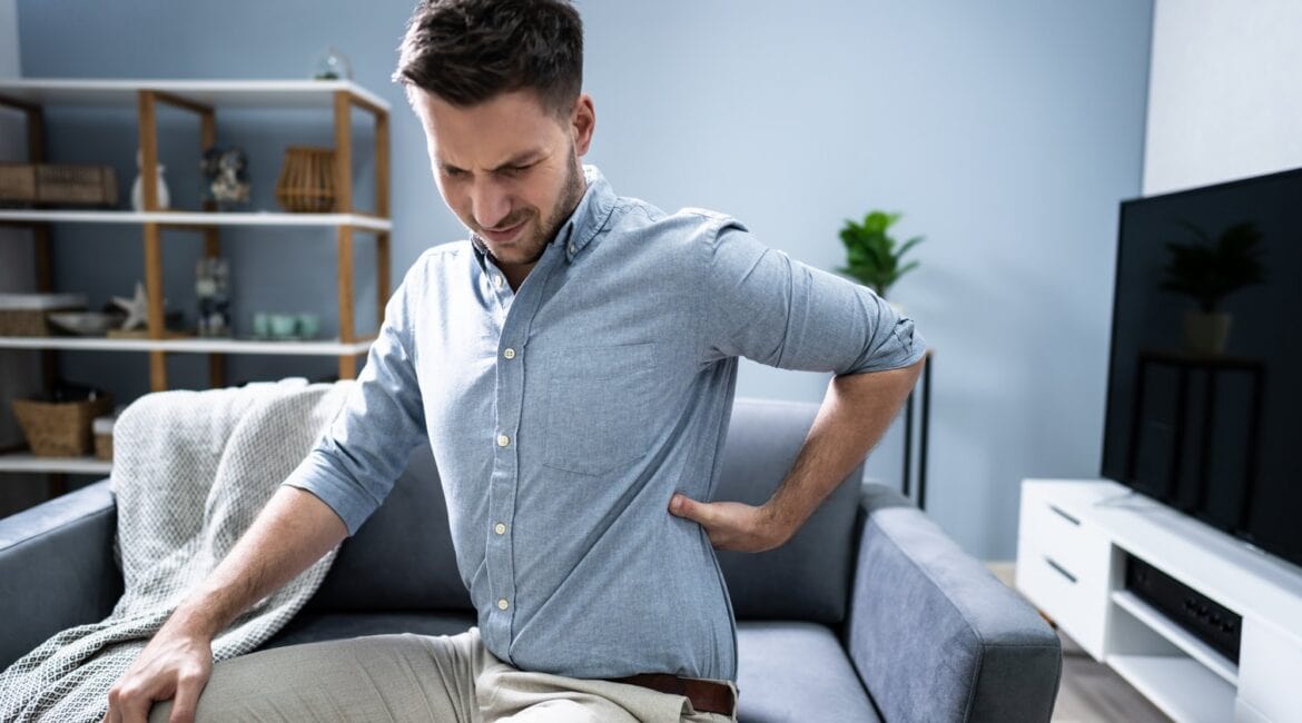 Man sitting on a sofa at home, holding his lower back and grimacing in pain