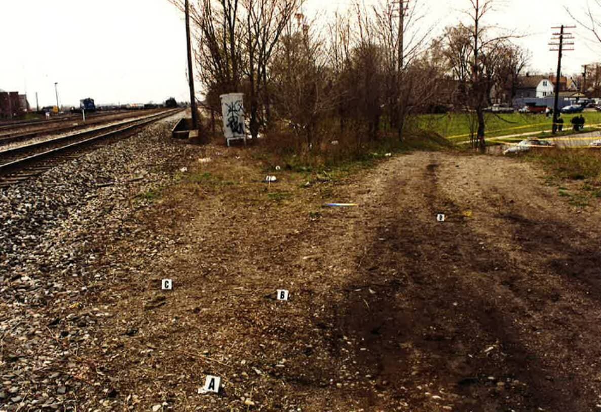 This view of the railroad track scene where the driveway leads up from the parking lot of a nearby building, looking west, showing how the service drive runs along the tracks. Image: DPD files.