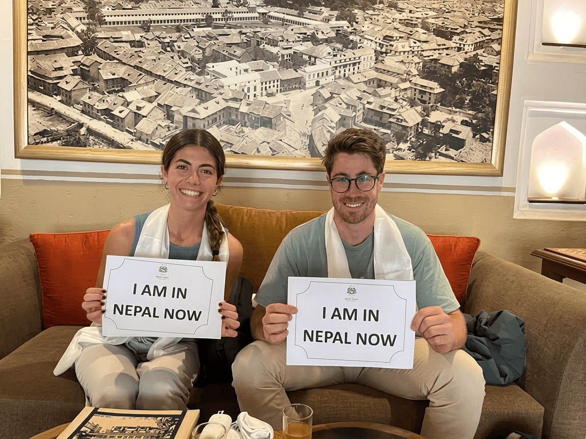 image A couple sitting on a couch and smiling while holding handmade signs that read "I AM IN NEPAL NOW," representing the tourists visiting the country.